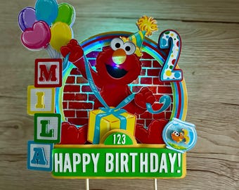 May include: A cake topper featuring a red Elmo character wearing a party hat, a yellow gift box, a number 2, and a fishbowl. The topper includes colorful balloons, blocks spelling MILA, and a green sign that reads HAPPY BIRTHDAY!