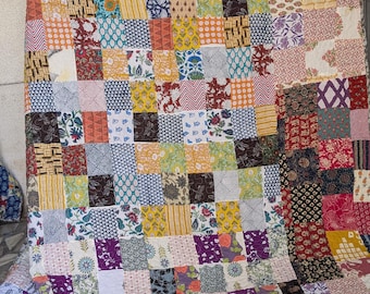 Puede incluir: Un edredón de retazos con una variedad de cuadrados de tela estampada en tonos de rojo, amarillo, azul, morado y verde. El edredón tiene una textura acolchada y un aspecto suave, ideal para una cama o un sofá.