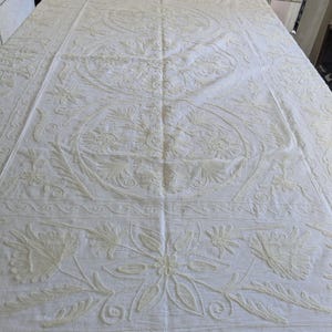 May include: Cream-colored embroidered tablecloth with floral and leaf patterns. The design features detailed stitching and a border with repeating motifs. The tablecloth is spread out, showcasing the intricate embroidery.