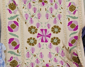 May include: Cream-coloured textile with detailed embroidery. It showcases a central floral design surrounded by repeating patterns of flowers, pomegranates, and leaves in shades of purple, green, and brown. The border has a decorative scroll design.