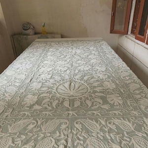 May include: A pale green bedspread with detailed white floral embroidery. The design includes a central floral pattern and repeating paisley-style motifs. The bedspread is laid out on a bed frame.