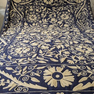 May include: A navy blue textile with intricate, cream-colored floral embroidery. The design features large, stylized flowers, swirling vines, and geometric borders, creating a rich, textured pattern across the fabric. The embroidery is detailed and visually striking.