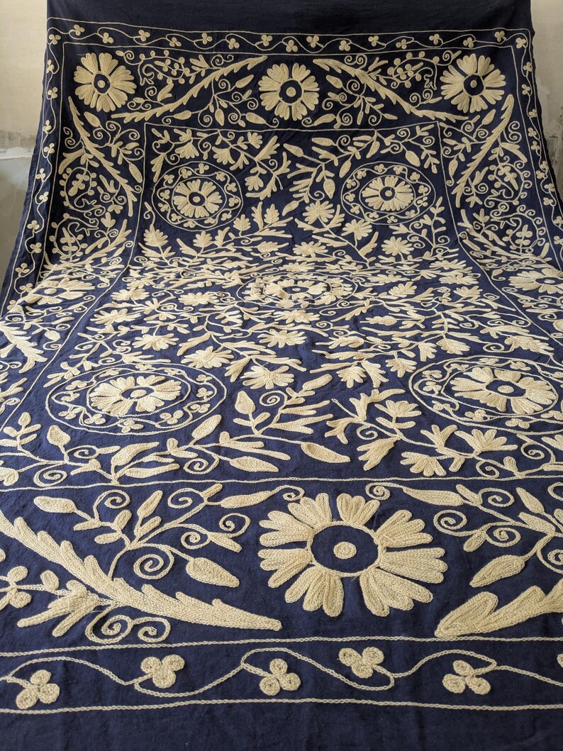 May include: A navy blue textile with intricate, cream-colored floral embroidery. The design features large, stylized flowers, swirling vines, and geometric borders, creating a rich, textured pattern across the fabric. The embroidery is detailed and visually striking.
