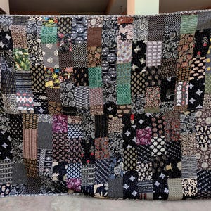 May include: A patchwork quilt featuring a variety of patterned fabrics in shades of black, brown, green, and purple. The quilt is made of numerous rectangular and square patches, creating a textured and visually rich design. The quilt is held up, showcasing its size and detail.
