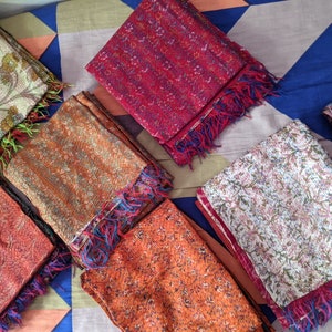 May include: A collection of colorful silk scarves with floral and paisley patterns. The scarves are folded and arranged on a patterned fabric background.