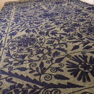 May include: A large, olive-green textile with intricate navy blue floral embroidery. The design features stylized flowers, leaves, and swirling patterns across the entire surface. The textile appears to be a blanket or a large decorative piece.
