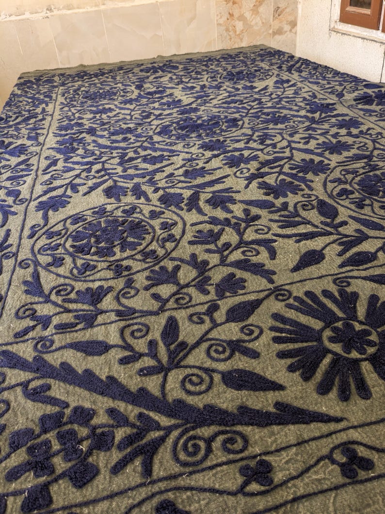 May include: A large, olive-green textile with intricate navy blue floral embroidery. The design features stylized flowers, leaves, and swirling patterns across the entire surface. The textile appears to be a blanket or a large decorative piece.