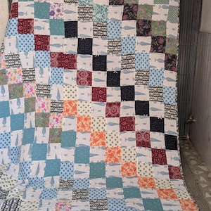 Puede incluir: Un edredón de retazos con una variedad de cuadrados estampados en tonos de azul, blanco, rojo, naranja y negro. Algunos cuadrados presentan motivos de ballenas. El edredón se muestra sobre un fondo neutro.