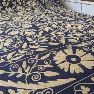 May include: A navy blue textile with intricate beige floral embroidery. The design features large flowers, leaves, and swirling patterns, creating a detailed and ornate appearance. The fabric is folded, revealing the pattern's repeating nature.