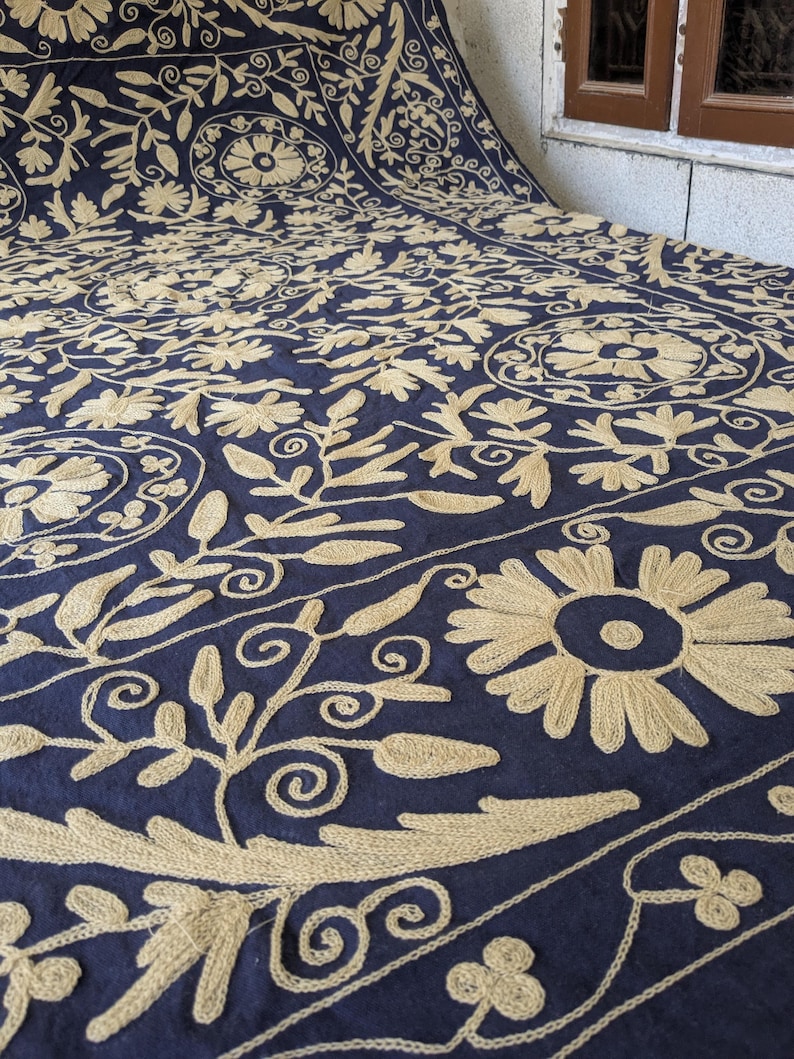 May include: A navy blue textile with intricate beige floral embroidery. The design features large flowers, leaves, and swirling patterns, creating a detailed and ornate appearance. The fabric is folded, revealing the pattern's repeating nature.