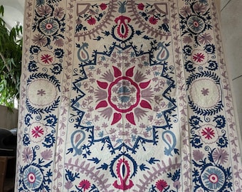May include: Cream-coloured textile with intricate embroidery. The design features a central floral medallion in shades of pink, blue, and purple, surrounded by detailed floral and geometric patterns. The edges are bordered with similar motifs.