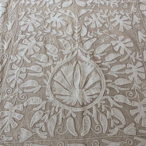 May include: Beige textile with intricate white floral embroidery. The design features stylized flowers, leaves, and swirling patterns. The embroidery is detailed and covers the entire surface, creating a textured and decorative effect. The textile appears to be a decorative item.