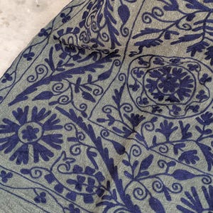 May include: Close-up of a textile with a detailed floral pattern. The fabric is a muted gray-green color, with intricate embroidery in a deep navy blue. The design features stylized flowers, leaves, and swirling vines.