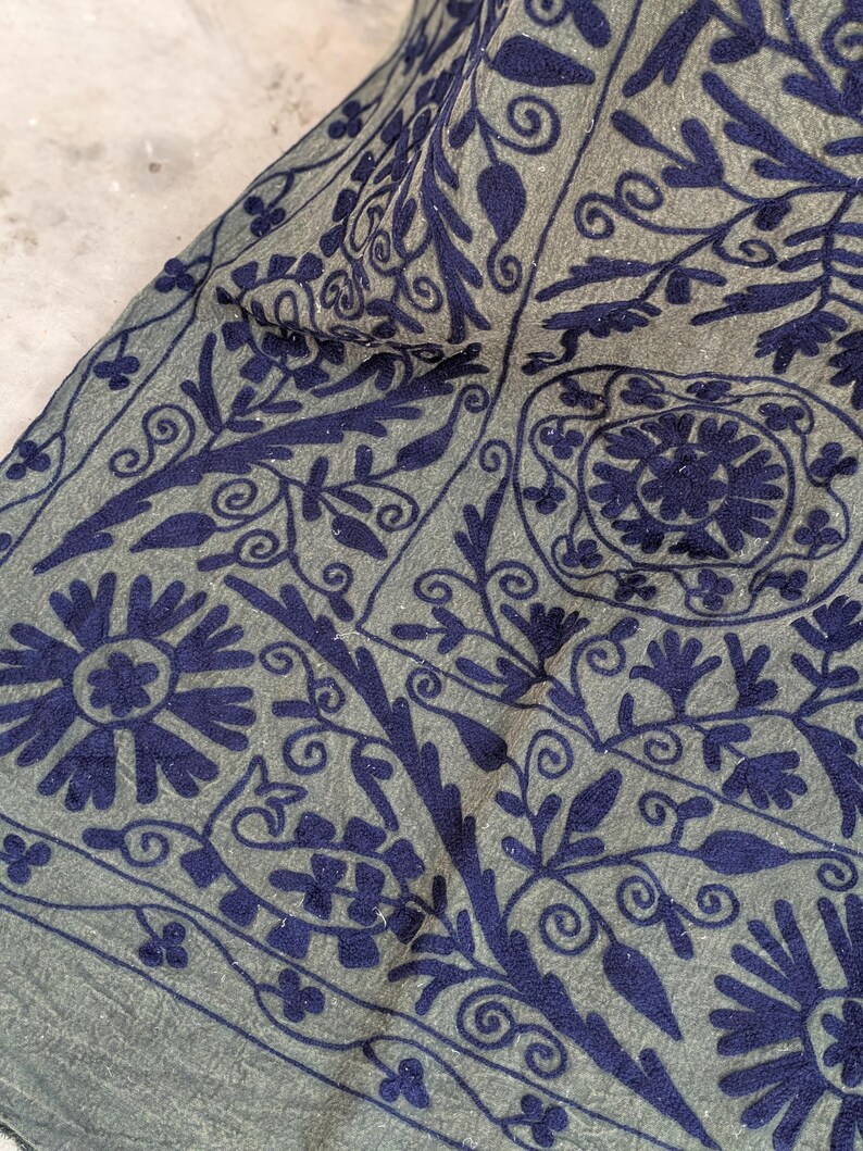 May include: Close-up of a textile with a detailed floral pattern. The fabric is a muted gray-green color, with intricate embroidery in a deep navy blue. The design features stylized flowers, leaves, and swirling vines.