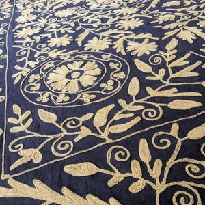 May include: A navy blue textile with intricate beige embroidery. The design features floral and leaf patterns, including stylized flowers and swirling vines. The embroidery is detailed and creates a textured surface.
