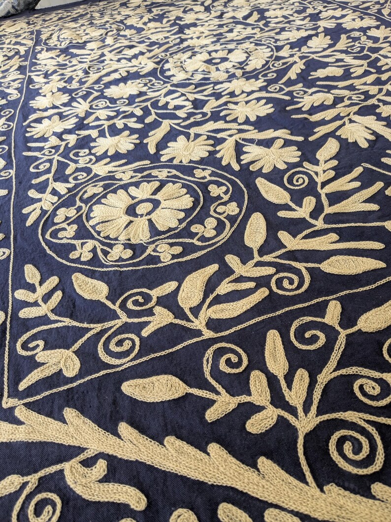 May include: A navy blue textile with intricate beige embroidery. The design features floral and leaf patterns, including stylized flowers and swirling vines. The embroidery is detailed and creates a textured surface.