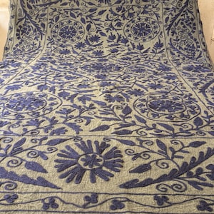 May include: A large, rectangular textile with a light gray background and intricate navy blue floral embroidery. The design features stylized flowers, leaves, and vines, creating a detailed pattern across the surface. The edges are finished with a decorative border.
