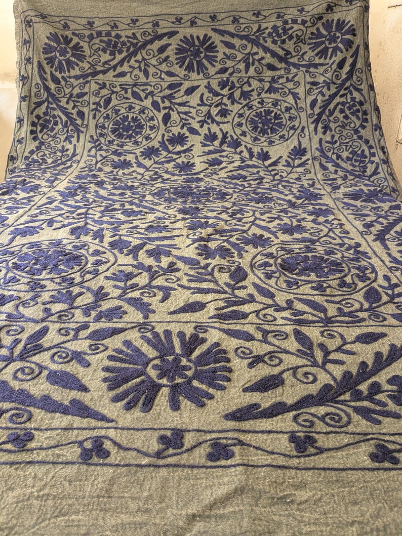 May include: A large, rectangular textile with a light gray background and intricate navy blue floral embroidery. The design features stylized flowers, leaves, and vines, creating a detailed pattern across the surface. The edges are finished with a decorative border.