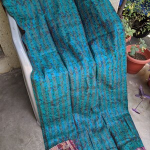 May include: A teal blue kantha quilt with a striped pattern and a colorful floral border. The quilt is made of recycled saris and has a fringed edge.