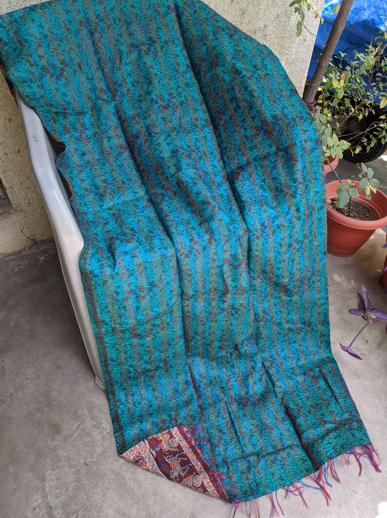 May include: A teal blue kantha quilt with a striped pattern and a colorful floral border. The quilt is made of recycled saris and has a fringed edge.