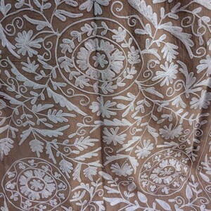 May include: Close-up of brown fabric with white floral embroidery. The design features detailed flowers, leaves, and swirling patterns, creating a textured surface. The embroidery is evenly distributed across the fabric.
