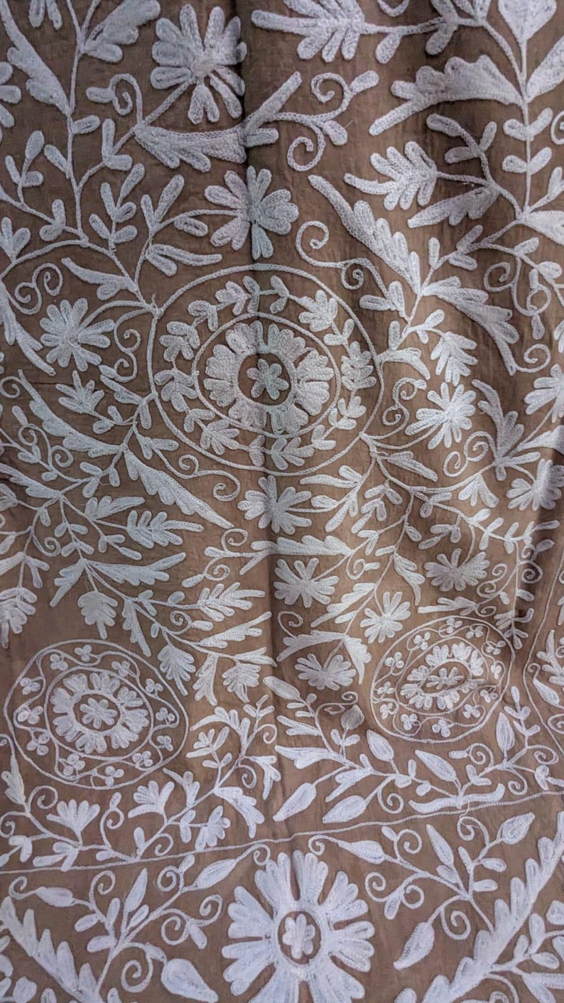 May include: Close-up of brown fabric with white floral embroidery. The design features detailed flowers, leaves, and swirling patterns, creating a textured surface. The embroidery is evenly distributed across the fabric.
