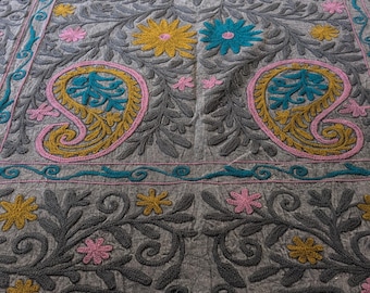May include: Gray textile with intricate floral embroidery. The design features yellow, pink, and teal flowers and paisley shapes. The edges are trimmed with a teal and pink border. The textile is likely a decorative item, such as a tablecloth or wall hanging.