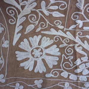 May include: Detailed shot of a brown fabric with white floral embroidery. The design includes large stylized flowers, curling vines, and delicate leaf patterns, creating a textured and detailed surface.