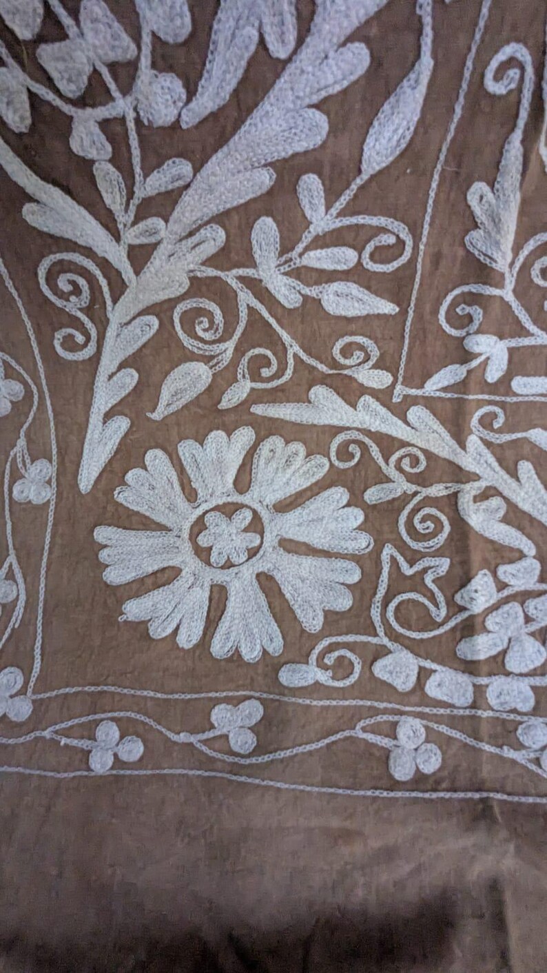 May include: Detailed shot of a brown fabric with white floral embroidery. The design includes large stylized flowers, curling vines, and delicate leaf patterns, creating a textured and detailed surface.