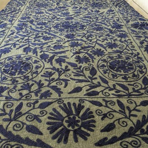 May include: A large, rectangular, olive green textile with an intricate embroidered floral pattern in navy blue. The design features stylized flowers, leaves, and vines, creating a detailed and ornate appearance. The textile is likely a decorative item, such as a throw or a rug.