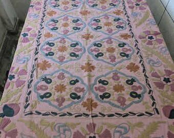 May include: A pink textile with intricate embroidery. The design features floral and geometric patterns in shades of pink, blue, green, and tan. The textile is likely a decorative item, such as a tablecloth or wall hanging.