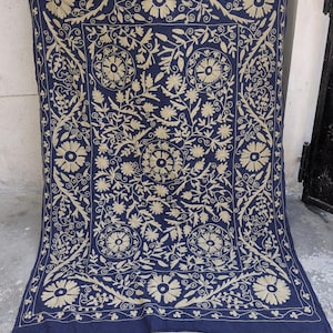 May include: A navy blue textile with intricate beige floral embroidery. The design features large, stylized flowers, leaves, and swirling patterns, creating a symmetrical, ornate composition. The edges are finished with a detailed border.