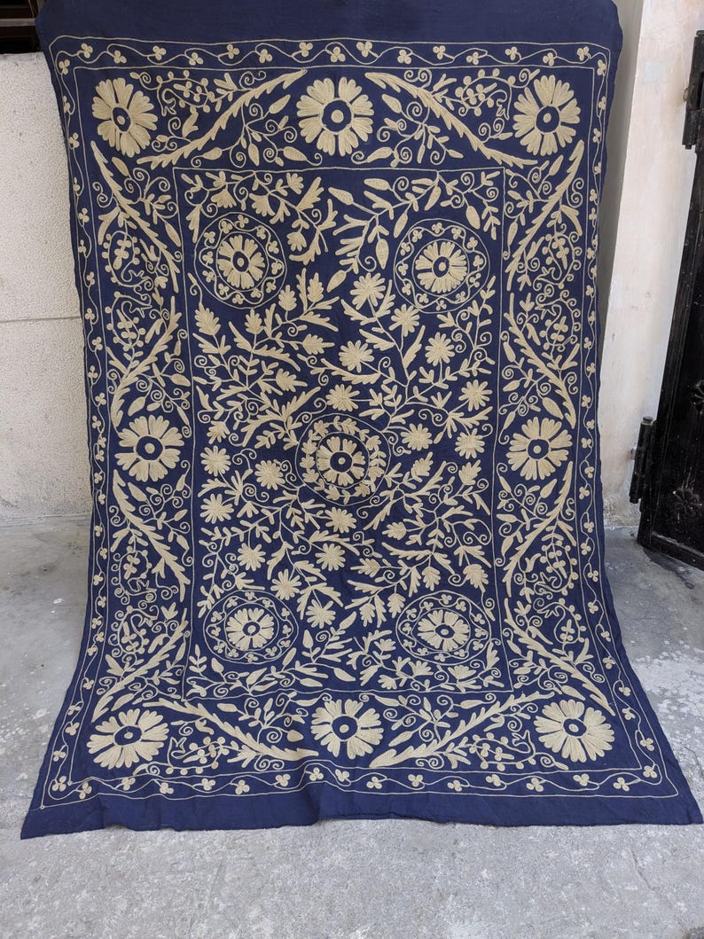 May include: A navy blue textile with intricate beige floral embroidery. The design features large, stylized flowers, leaves, and swirling patterns, creating a symmetrical, ornate composition. The edges are finished with a detailed border.