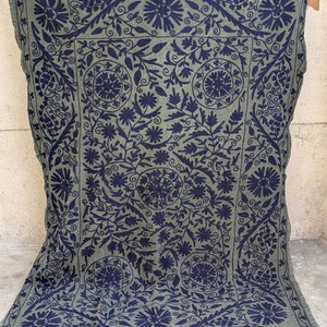 May include: A rectangular, olive green textile with a navy blue floral pattern. The design features stylized flowers, leaves, and swirling vines, creating a symmetrical, ornate border and central medallion. The textile is likely a blanket or throw.