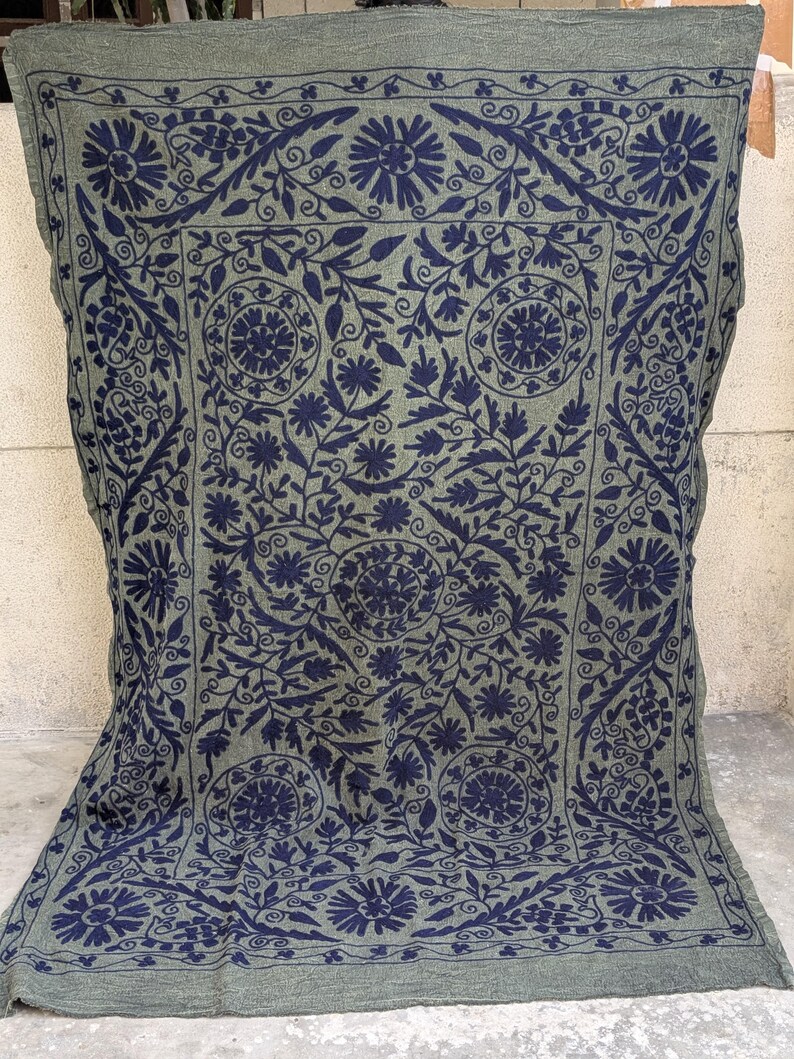 May include: A rectangular, olive green textile with a navy blue floral pattern. The design features stylized flowers, leaves, and swirling vines, creating a symmetrical, ornate border and central medallion. The textile is likely a blanket or throw.