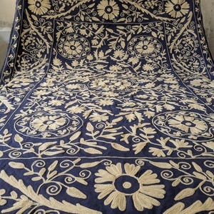 May include: A navy blue textile with intricate beige floral embroidery. The design features large flowers, swirling vines, and geometric patterns, creating a detailed and ornate appearance. The textile is likely a decorative piece, such as a throw or wall hanging.