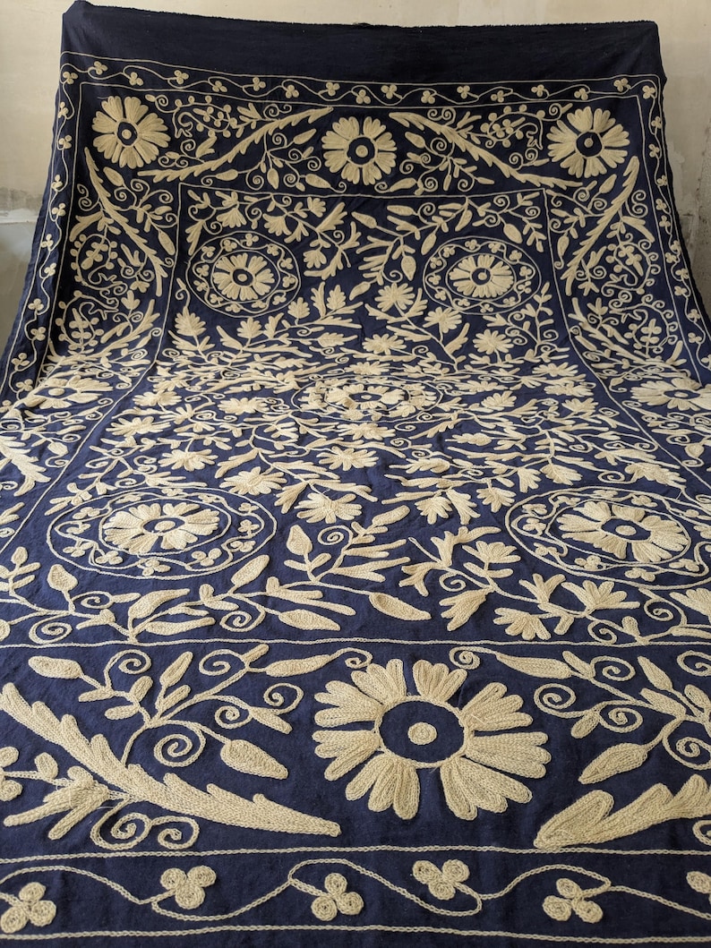 May include: A navy blue textile with intricate beige floral embroidery. The design features large flowers, swirling vines, and geometric patterns, creating a detailed and ornate appearance. The textile is likely a decorative piece, such as a throw or wall hanging.