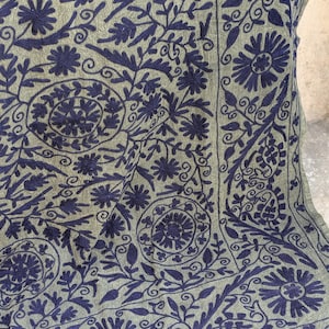 May include: Close-up of a green textile with intricate dark blue floral embroidery. The design features stylized flowers, leaves, and swirling patterns, creating a detailed and textured surface. The embroidery is densely packed, covering the fabric.