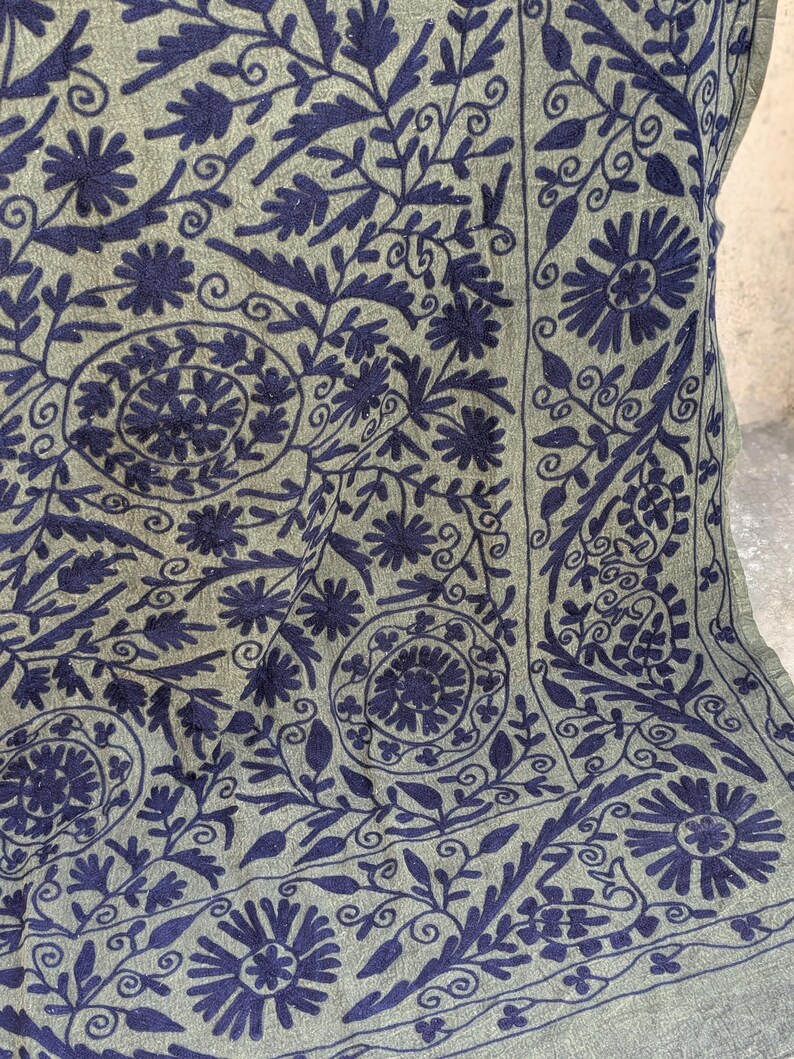 May include: Close-up of a green textile with intricate dark blue floral embroidery. The design features stylized flowers, leaves, and swirling patterns, creating a detailed and textured surface. The embroidery is densely packed, covering the fabric.