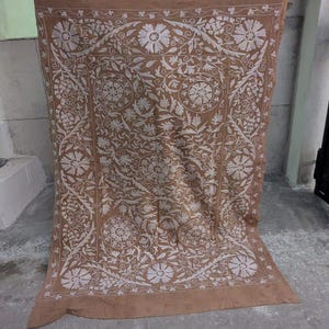 May include: A brown textile with a detailed white floral pattern. The design features large, stylized flowers and scrolling vines, creating a symmetrical and ornate composition. The textile is likely a decorative piece, such as a tapestry or wall hanging.