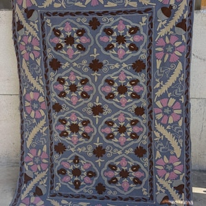Op de afbeelding: Een rechthoekig, blauw textiel met een ingewikkeld bloemmotief. De borduursels bevatten roze, bruine en beige bloemen en bladeren. De rand heeft een vergelijkbaar bloemmotief. Het textiel hangt tegen een lichtgekleurde muur.