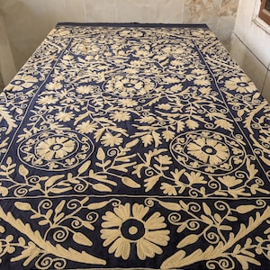 May include: A navy blue textile with intricate gold floral embroidery. The design features large, stylized flowers, leaves, and swirling patterns. The embroidered details create a rich, textured appearance, suitable for a decorative textile or a table covering.