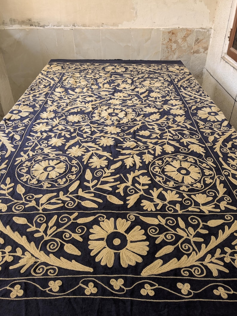 May include: A navy blue textile with intricate gold floral embroidery. The design features large, stylized flowers, leaves, and swirling patterns. The embroidered details create a rich, textured appearance, suitable for a decorative textile or a table covering.