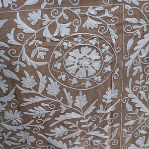 May include: Close-up of a brown textile with detailed white floral embroidery. The design features leaves, flowers, and swirling patterns, creating a textured and ornate appearance. The embroidery is evenly spaced across the fabric.