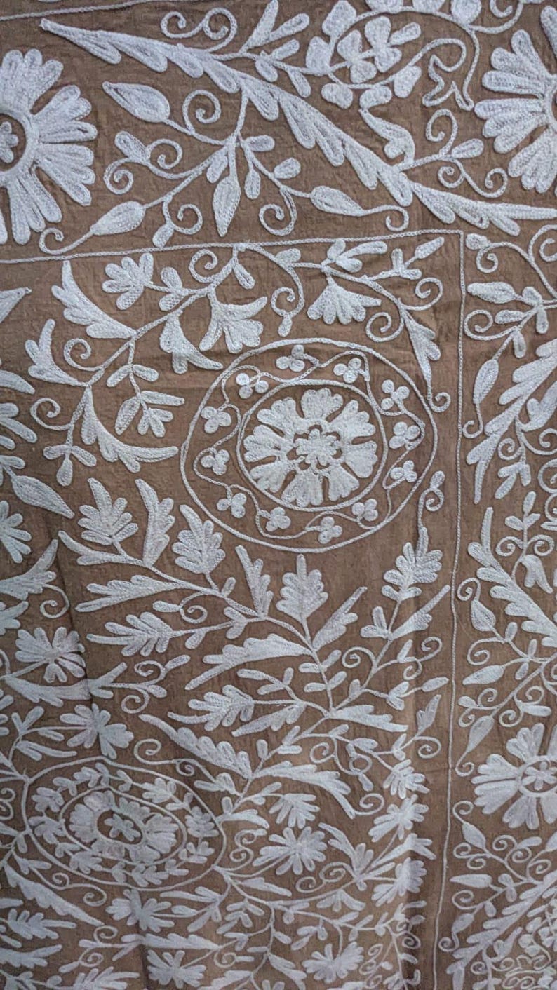 May include: Close-up of a brown textile with detailed white floral embroidery. The design features leaves, flowers, and swirling patterns, creating a textured and ornate appearance. The embroidery is evenly spaced across the fabric.