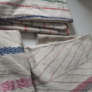 Op de afbeelding: Een verzameling vintage katoenen handdoeken met een getextureerde afwerking. De handdoeken zijn voornamelijk gebroken wit met gekleurde strepen in rood, groen en blauw. Sommige hebben roze en grijze strepen of decoratieve blauwe en roze patronen.