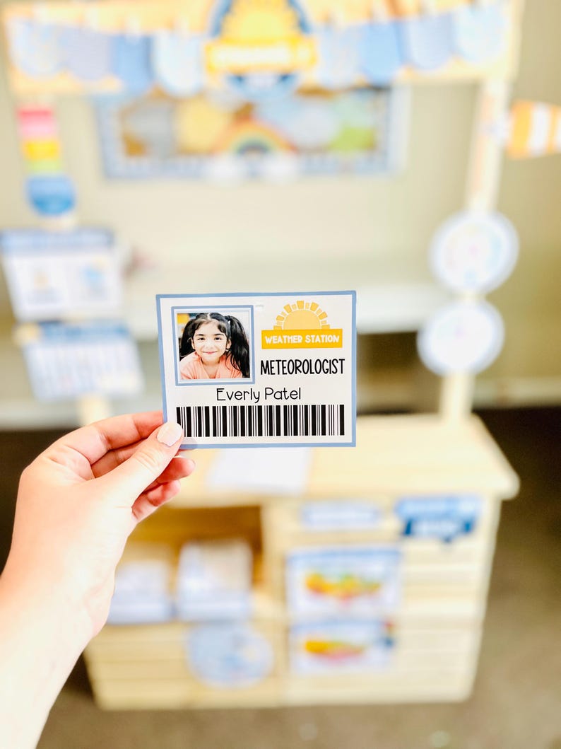 May include: A child's weather station play set with a name tag that reads "Weather Station Meteorologist Everly Patel". The play set is made of light-colored wood and has a blue and yellow awning.
