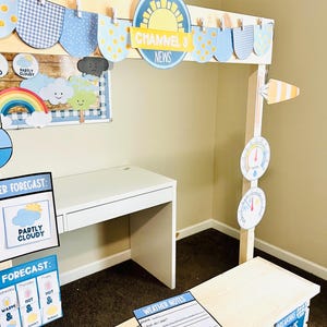 May include: A children's weather station play set with a white desk, a wooden news desk, and various weather-themed decorations. The set includes signs with the text "Channel 3 News", "Weather Forecast", and "Breaking News".