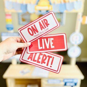 May include: Three red and white rectangular signs with text: "ON AIR Please Stay Quiet", "LIVE", and "ALERT!". The signs are held in front of a wooden play stand with a blue and yellow awning.