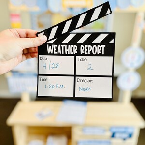 May include: A black and white clapperboard with the text "WEATHER REPORT" and handwritten details: date 4/28, take 2, time 1:20 P.M., and director Noah. The clapperboard is held in front of a wooden play stand.